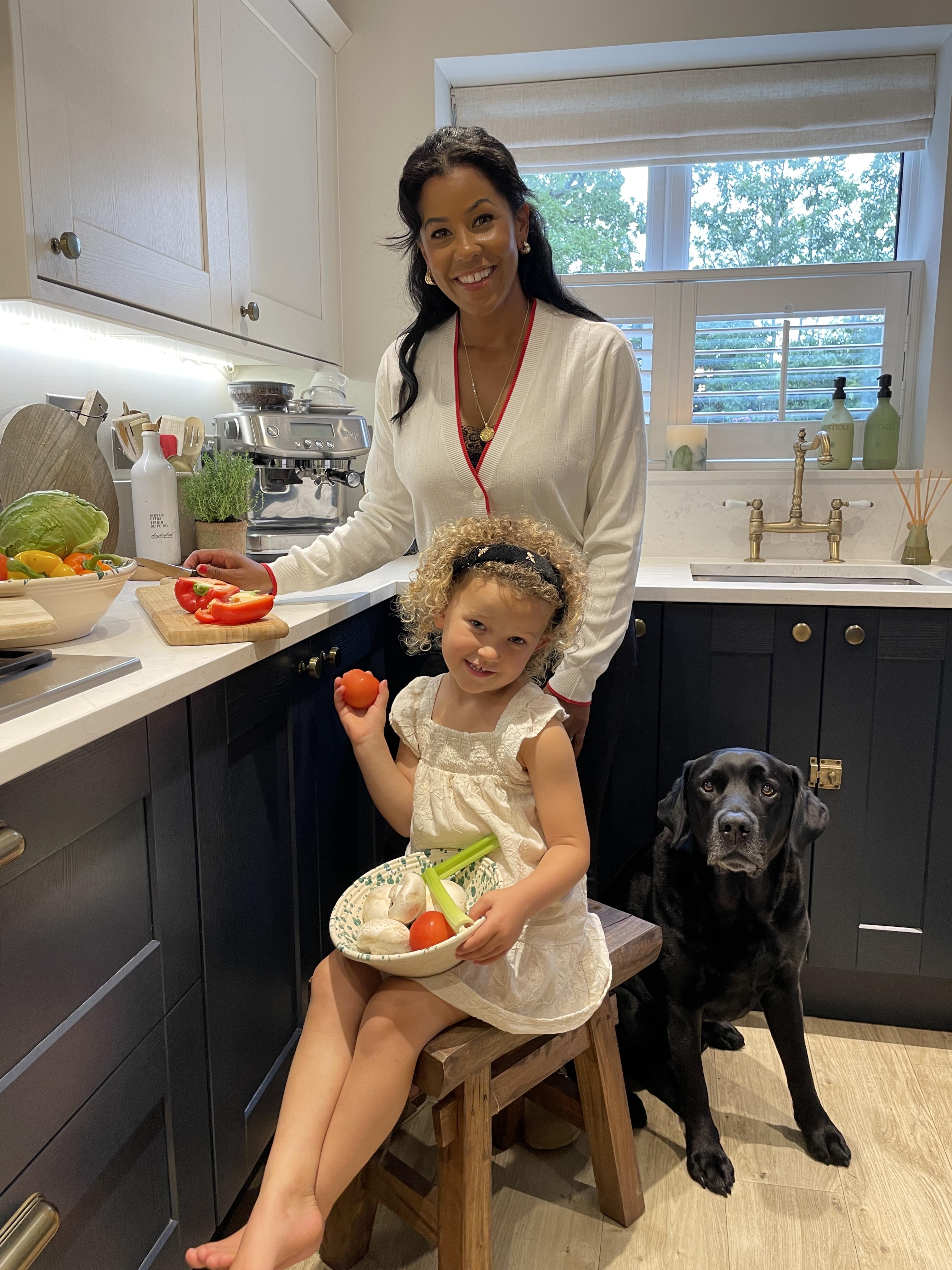 A woman, a little girl, and a black dog in a kitchen. They are holding vegetables.