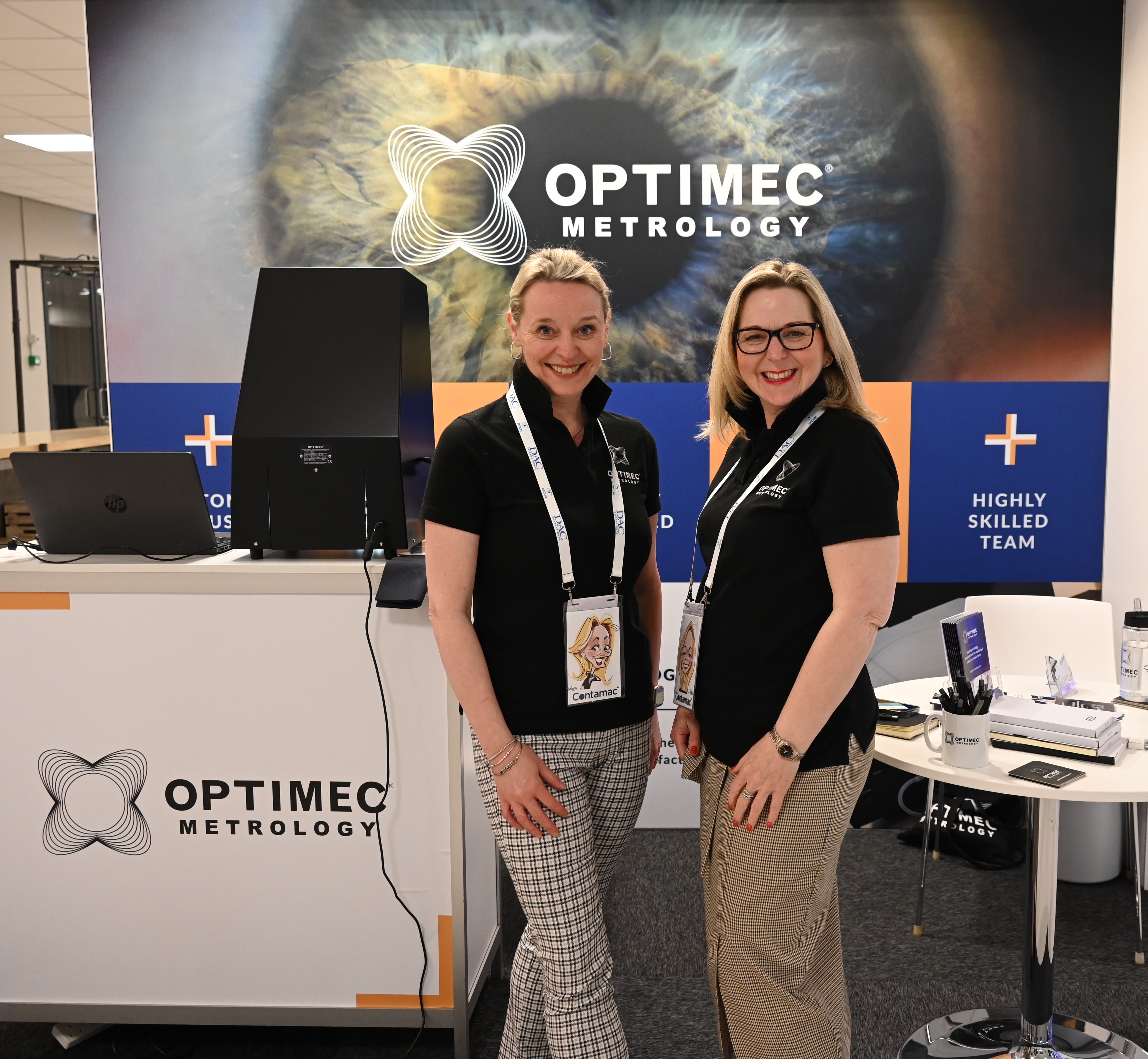 two members of staff standing by the optimec metrology show stand